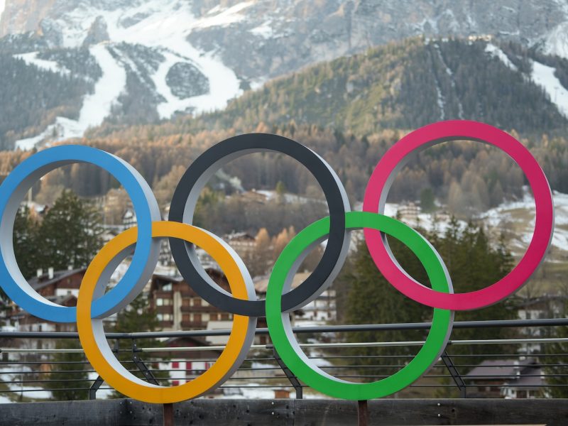 Colorful Olympic rings displayed outdoors with snow‑covered mountains in the background.