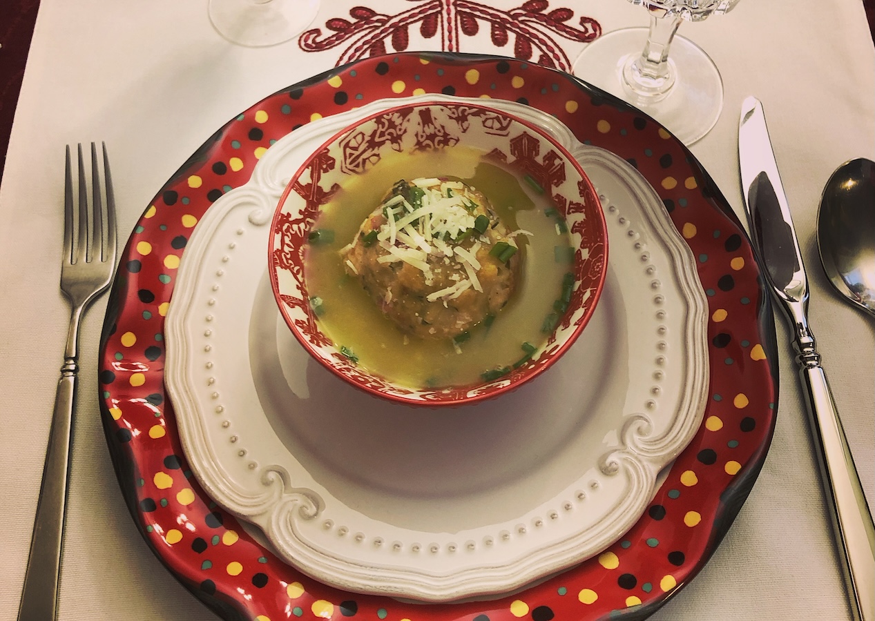 A small bowl of soup with a dumpling and grated cheese, placed on decorative layered plates with cutlery and wine glasses.