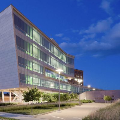 Contemporary glass-facade building illuminated at twilight with green interior lighting, surrounded by landscaped grounds and walkway.