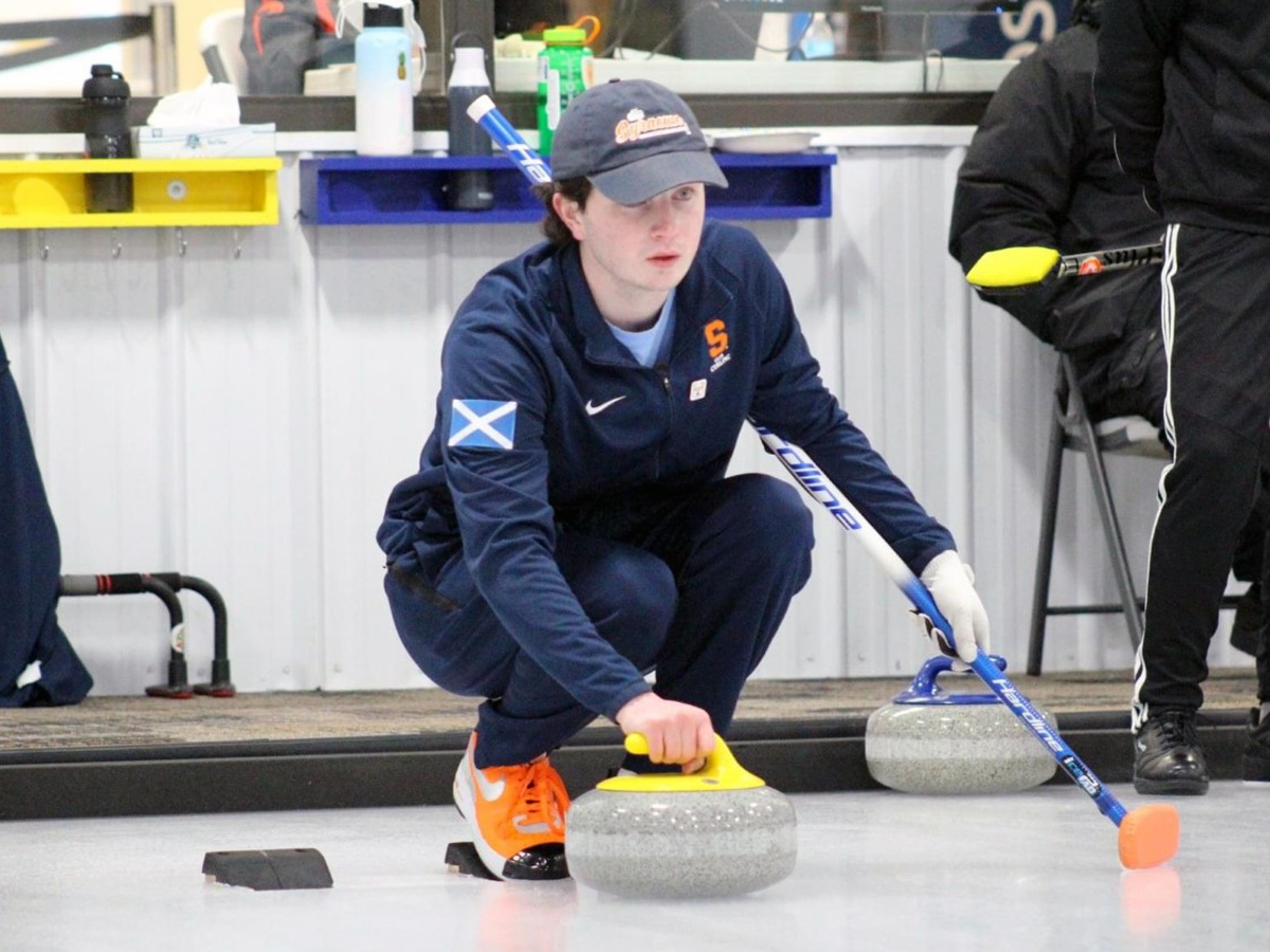 A Syracuse University curler in navy uniform crouches on the ice, hand on the stone's handle, ready to deliver.