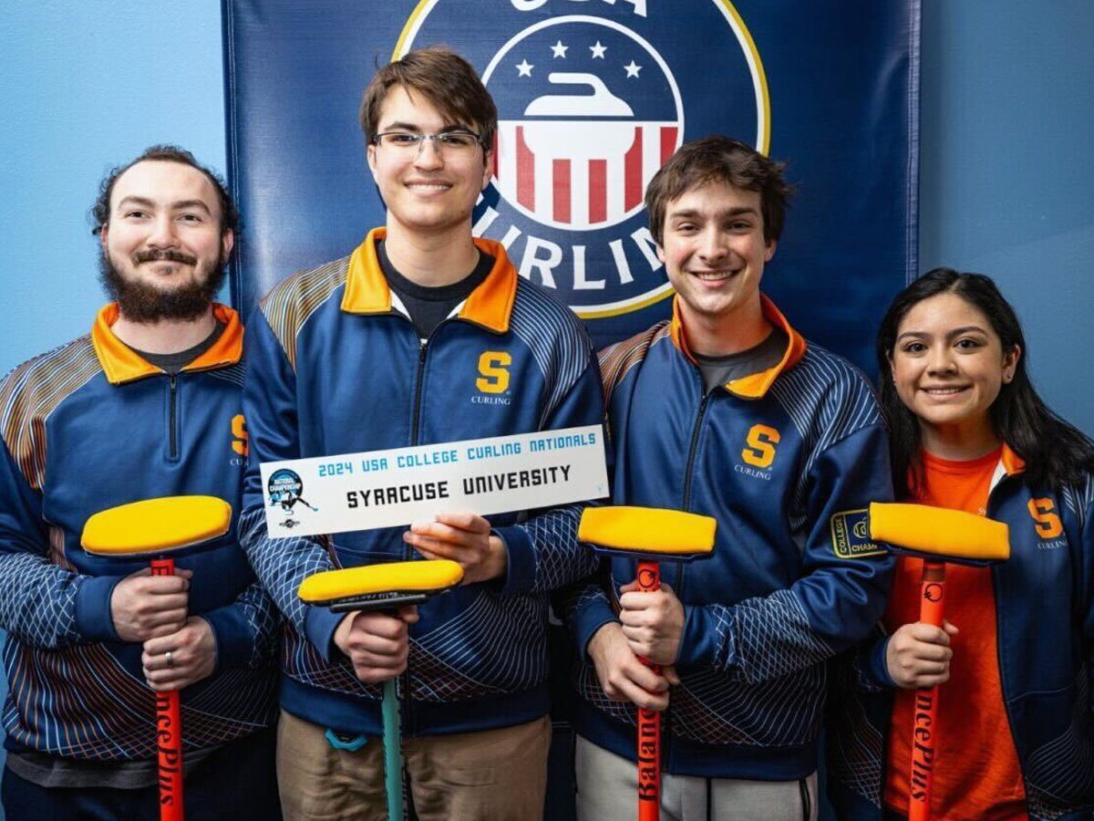 Four Syracuse University curling team members pose with their brooms and a sign reading "2024 USA College Curling Nationals Syracuse University.