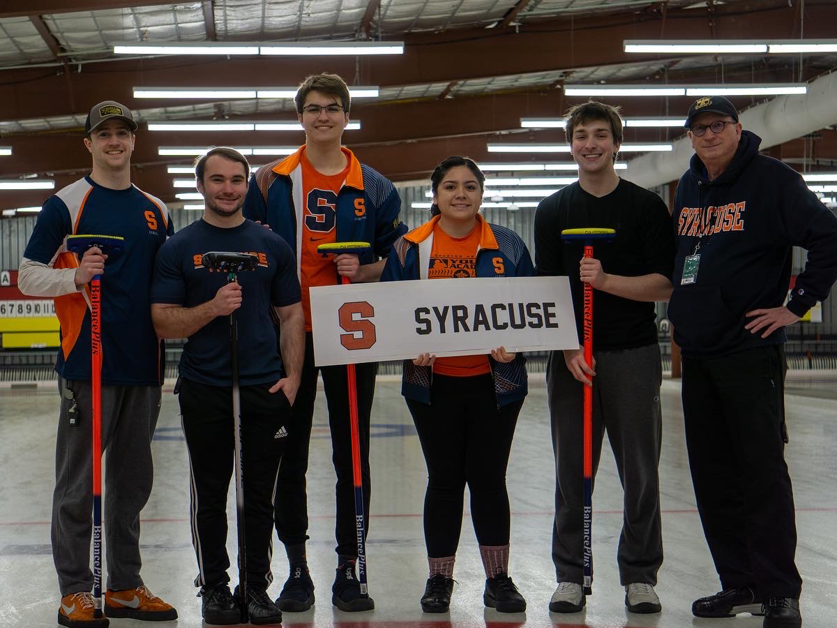 The Syracuse University curling team stands on the ice holding brooms and a Syracuse banner.