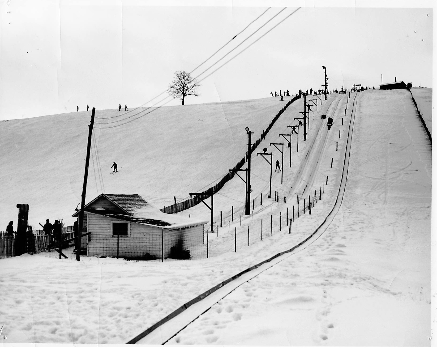 Skiers on a snowy hill with a tow lift and a small shack at the base.