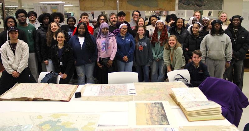 Large group of students standing together in a library or archives room behind several large historical maps spread out on a table.