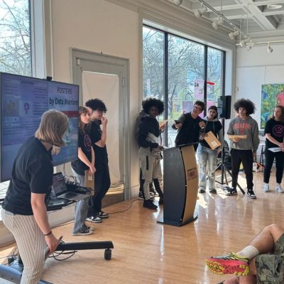 A group of young people presenting "Posters by Data Warriors" at a podium in a bright, art-filled community space, with a research poster displayed on a large screen beside them and audience members seated nearby.