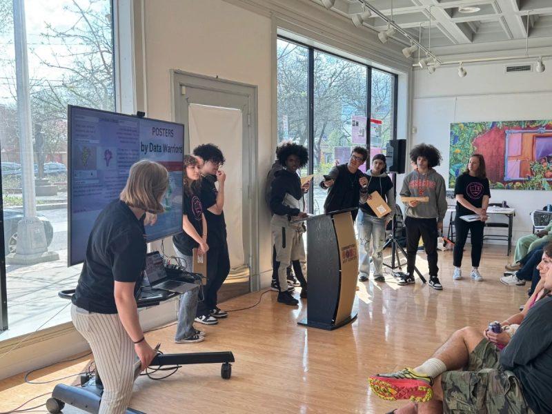 A group of young people presenting "Posters by Data Warriors" at a podium in a bright, art-filled community space, with a research poster displayed on a large screen beside them and audience members seated nearby.