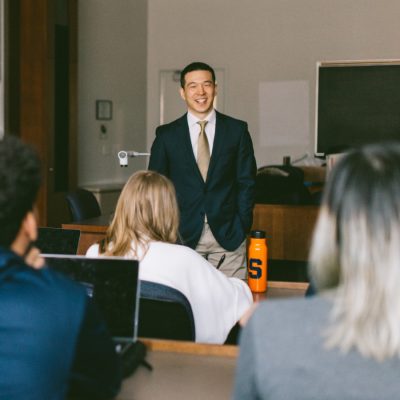Person in a suit standing at the front of a classroom while students listen from their desk