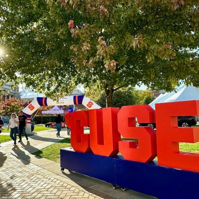 Large red "'CUSE" block letters on a blue base on a sunny Syracuse University campus lawn during an outdoor event.
