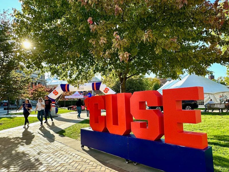 Large red "'CUSE" block letters on a blue base on a sunny Syracuse University campus lawn during an outdoor event.