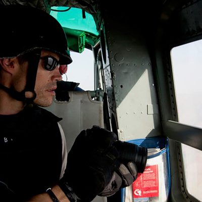 A helmeted photographer holds a camera while looking out the window of a military helicopter