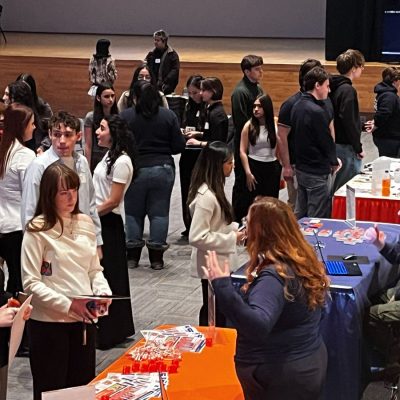 A group of students mingles with representatives seated at tables draped in orange and blue during an indoor event in a large hall. Brochures and promotional materials are displayed on the tables.