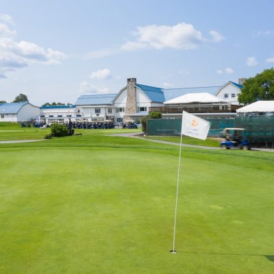 Golf green with a flag in the foreground and a clubhouse with blue roofs in the background under a partly cloudy sky.