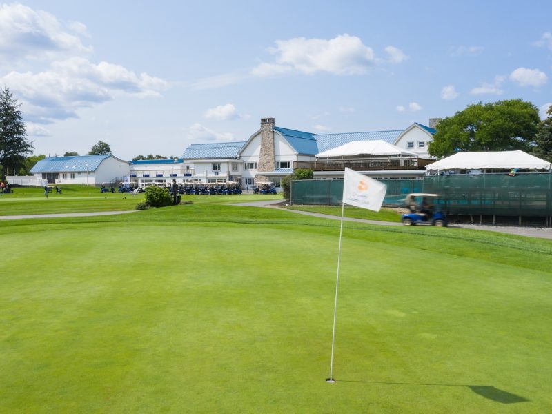 Golf green with a flag in the foreground and a clubhouse with blue roofs in the background under a partly cloudy sky.