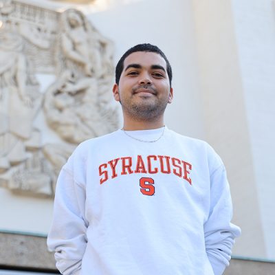 Person wearing a Syracuse sweatshirt standing outdoors in front of a building with architectural artwork