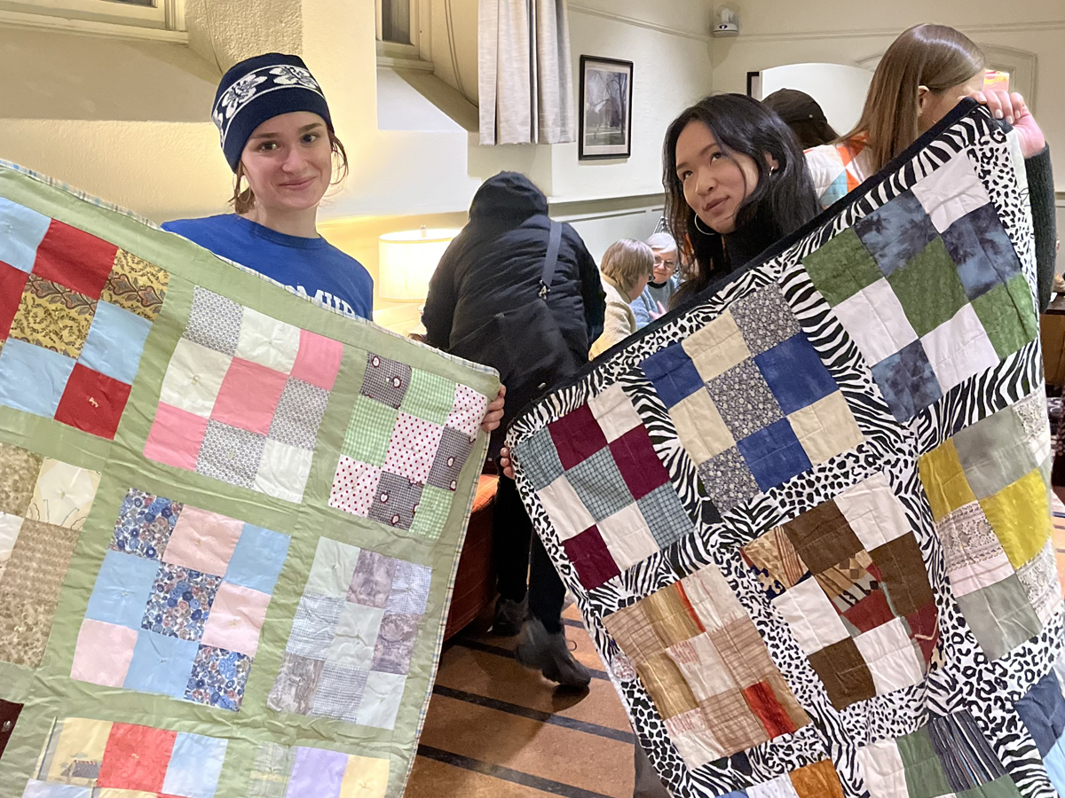 Two students hold up handmade patchwork quilts — one with a soft green border and pastel squares, the other with a bold black-and-white animal print border and colorful patches.
