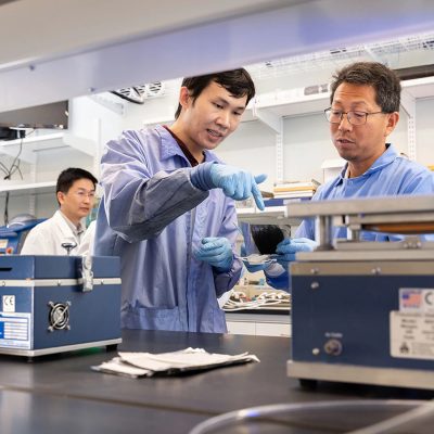 Two men in blue coats work with equipment in a lab. A third man in a white coat appears in the background.