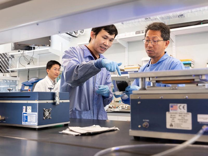 Two men in blue coats work with equipment in a lab. A third man in a white coat appears in the background.