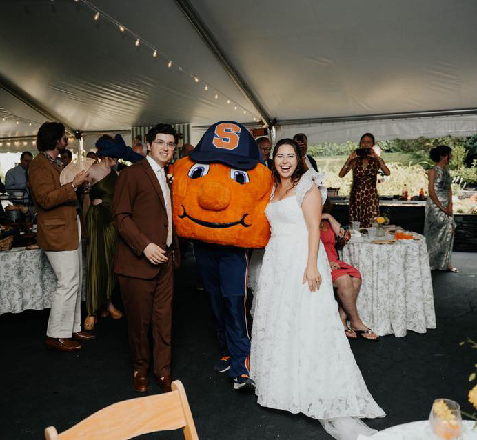 Bride and groom posing with Otto the Orange mascot at a wedding reception under a large tent, with guests celebrating in the background