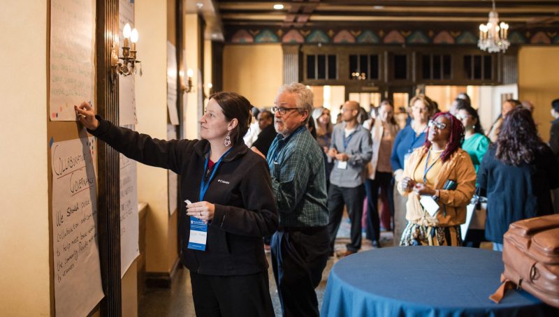 A woman points to notes on a large paper posted on a wall while a group of conference attendees looks on.
