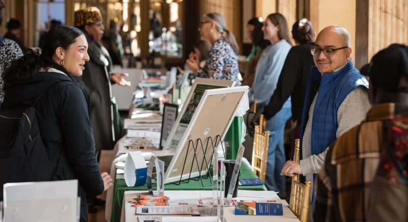 A staff member speaks with an attendee across a table at an indoor resource fair or information event, with additional tables and attendees visible in the background.