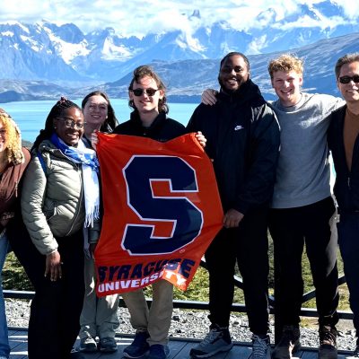 A group of seven people stand together outdoors holding a Syracuse University orange flag with the blue "S" logo. Behind them is a scenic backdrop of snow-capped mountains and a blue lake. The group is dressed in casual outdoor clothing and jackets.