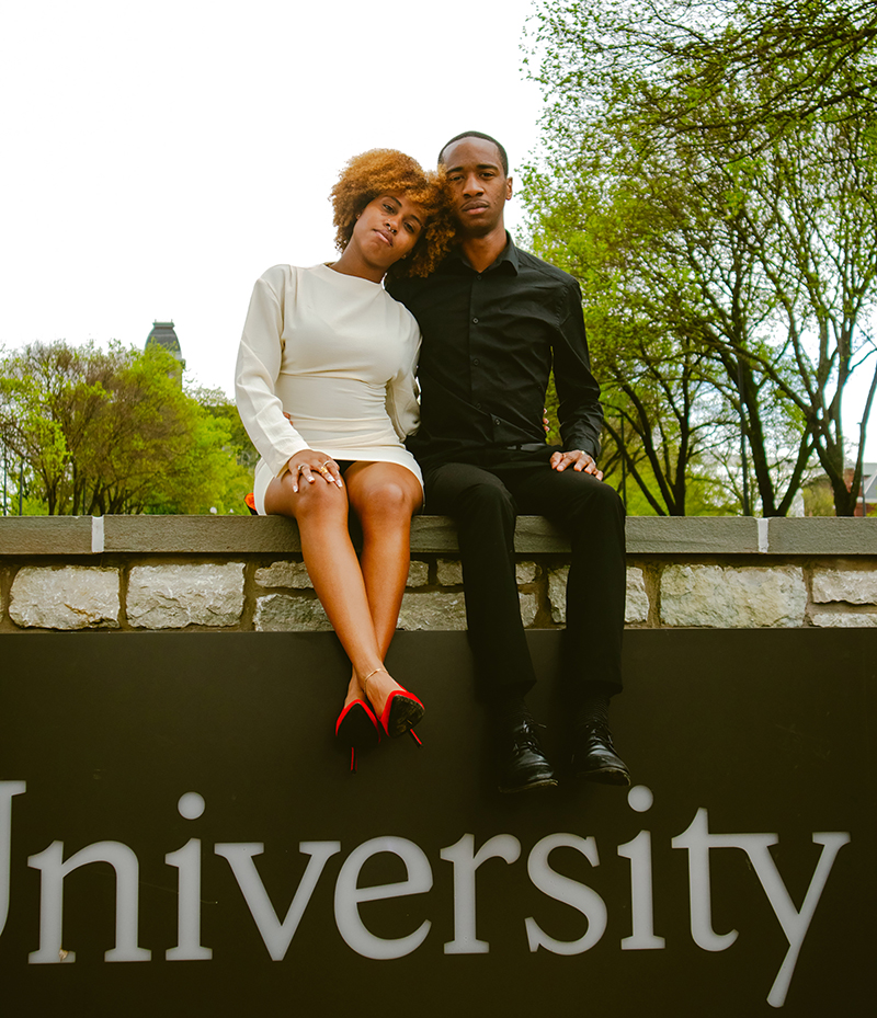 Two people dressed in formal attire sitting together on a stone ledge in front of a university sign, surrounded by spring trees