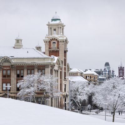 A snow-covered Syracuse University campus in winter, featuring the ornate red brick and cream-trimmed facade of Lyman Hall surrounded by snow-dusted trees and a snow-blanketed hillside under an overcast gray sky.