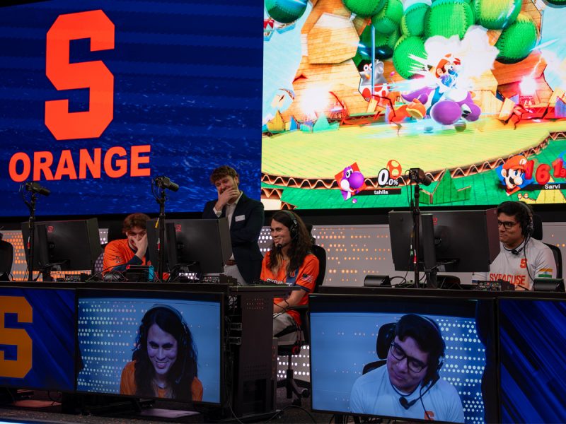An esports team competes on stage at individual gaming stations, with a large screen above showing live gameplay and bold orange team branding across the arena backdrop.