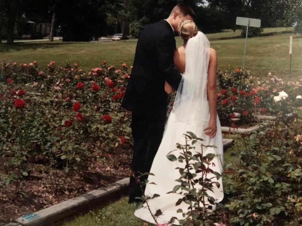 Bride and groom standing together in a garden filled with blooming roses, with the bride’s veil flowing behind her
