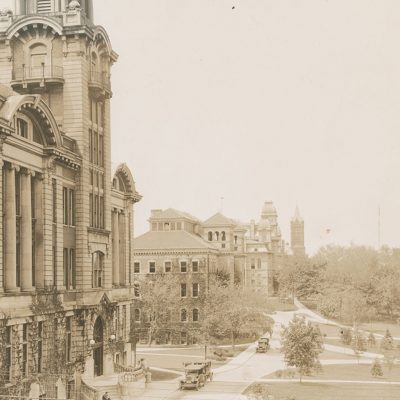 Historic campus buildings line a gently curving walkway, with trees, lawns, and a few early automobiles in the foreground.