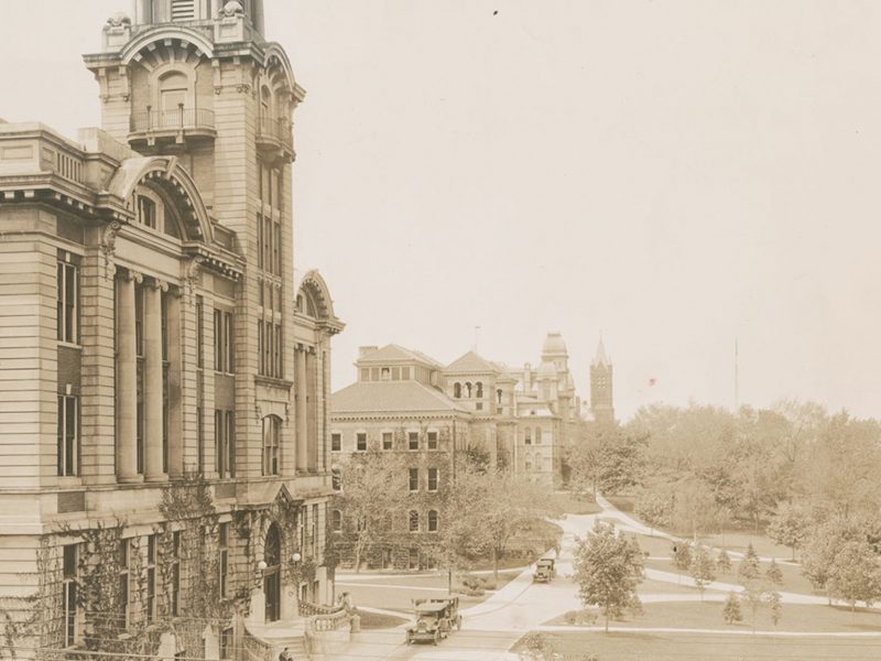 Historic campus buildings line a gently curving walkway, with trees, lawns, and a few early automobiles in the foreground.
