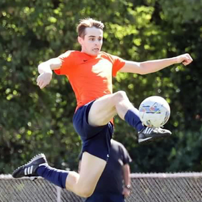 एक व्यक्ति नारंगी जर्सी टॉप और नीली शॉर्ट्स पहने हुए फुटबॉल की गेंद को ड्रिबल कर रहा है।