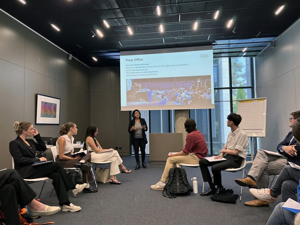 Woman presents Olympic Press Office information to group of seated students in contemporary classroom setting with projection screen.