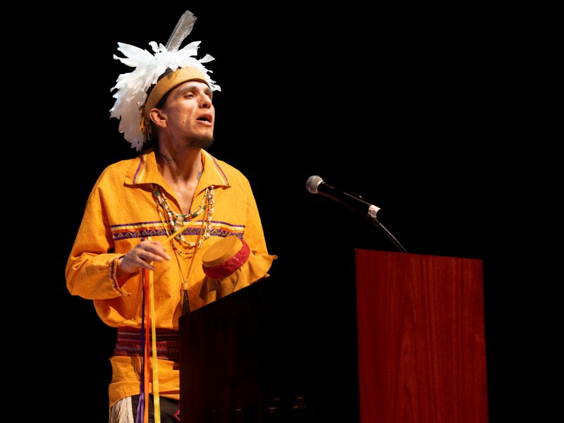 A man in traditional Indigenous regalia, including a white feathered headdress and yellow embroidered shirt, speaks at a podium on a darkened stage.