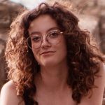 Headshot of a person with curly auburn hair and round glasses, smiling slightly, photographed outdoors in natural light.