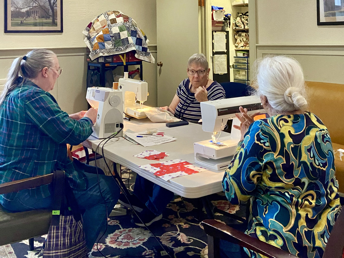 Three women sit around a table in a cozy room, working at sewing machines on colorful quilts.