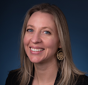 A person with long light‑colored hair wearing a dark blazer and decorative earrings against a dark background.