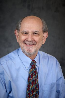 Person wearing a light blue dress shirt and patterned tie in front of a dark studio backdrop