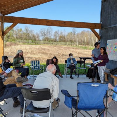 A small group gathers in an open-air pavilion overlooking farmland for an outdoor meeting with a sticky note board.