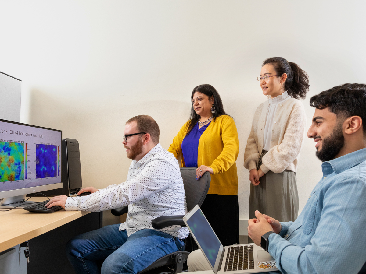 A professor stands with three student researchers gathered around a desktop computer displaying molecular simulation data in a lab setting.