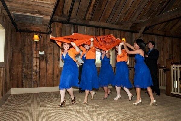 Bridesmaids in blue dresses and orange sweaters joyfully holding a large orange Syracuse banner overhead as they walk together inside a rustic wooden venue