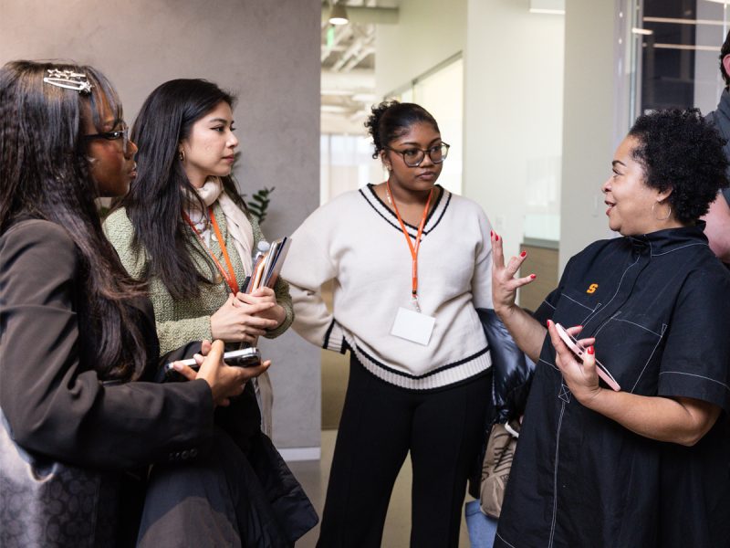 Three students with orange lanyards engage in conversation with a professional at a networking event.