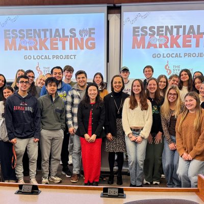 Group of approximately 40 marketing students posing for a class photo in a university lecture hall, with two projection screens displaying 'Essentials of Marketing: Go Local Project' in the background.