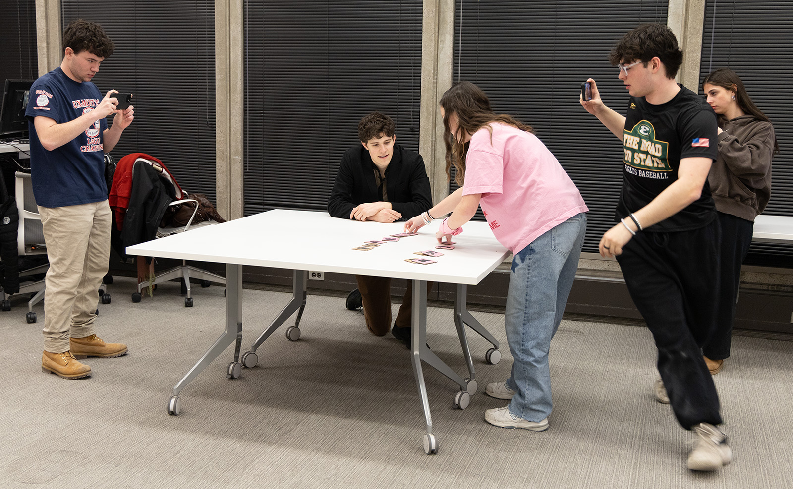 Several people gather around a table as one participant arranges cards while others record the activity with their phones