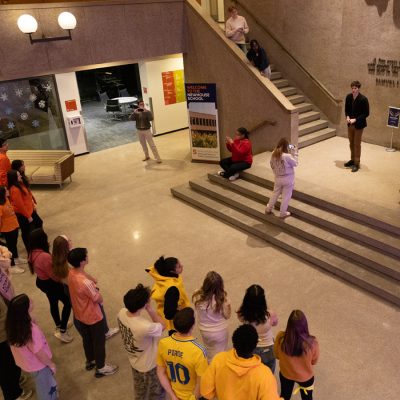 Students gather in a campus building atrium, watching participants on a staircase film the premiere episode of a "Survivor"-style competition