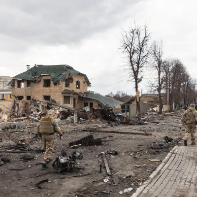 Two Ukrainian soldiers walk down a debris-strewn street lined with destroyed buildings and damaged trees, surveying the aftermath of heavy fighting in a residential area.