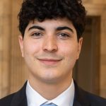 Headshot of a young person with curly dark hair, smiling, wearing a black suit jacket, white dress shirt, and light blue tie, photographed in front of a building.