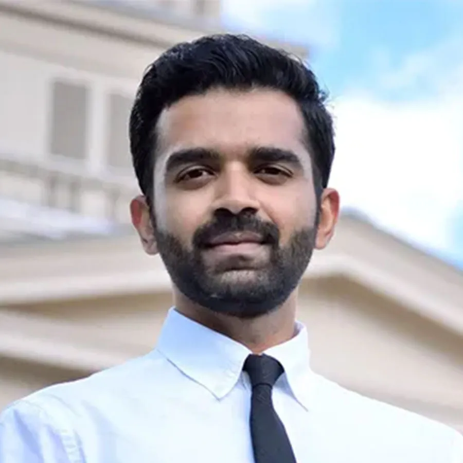 Person wearing a white dress shirt and black tie, standing outdoors in front of a light-colored building under a blue sky