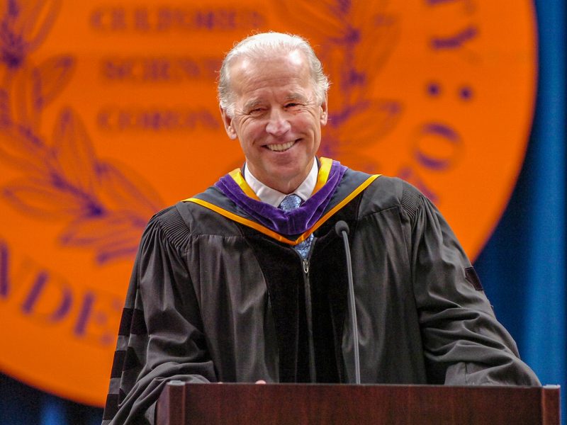 A person wearing black academic regalia with a purple and gold hood stands at a podium during a commencement ceremony, with an orange university seal visible in the background