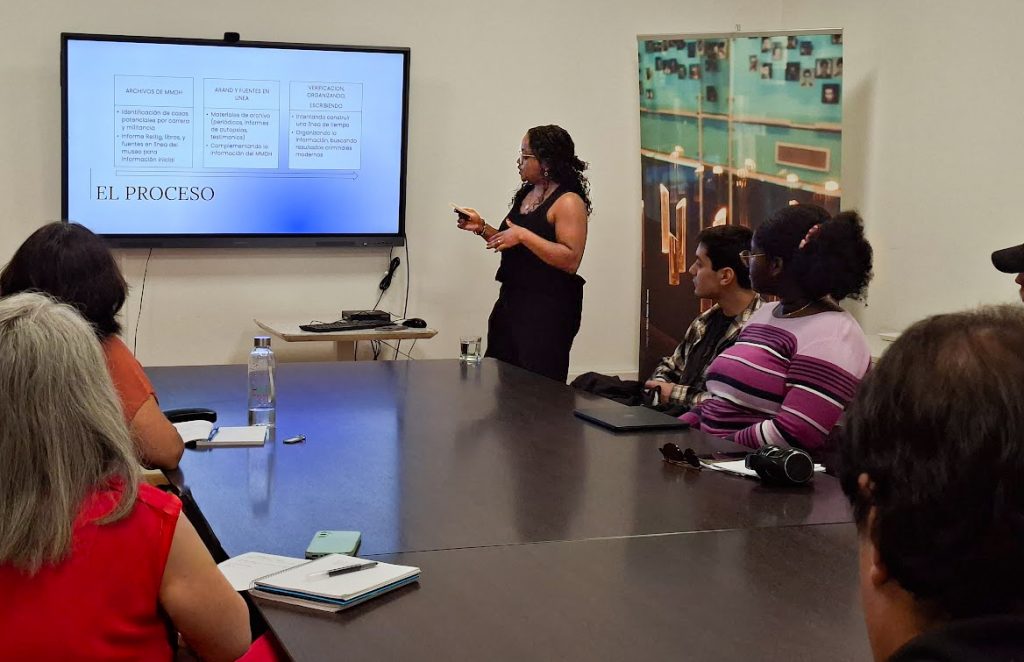 A woman stands at the front of a conference room presenting to a seated audience. Behind her, a large screen displays a presentation slide titled "EL PROCESO" with text in Spanish. Several attendees sit around a dark conference table, with artwork visible on the wall in the background.